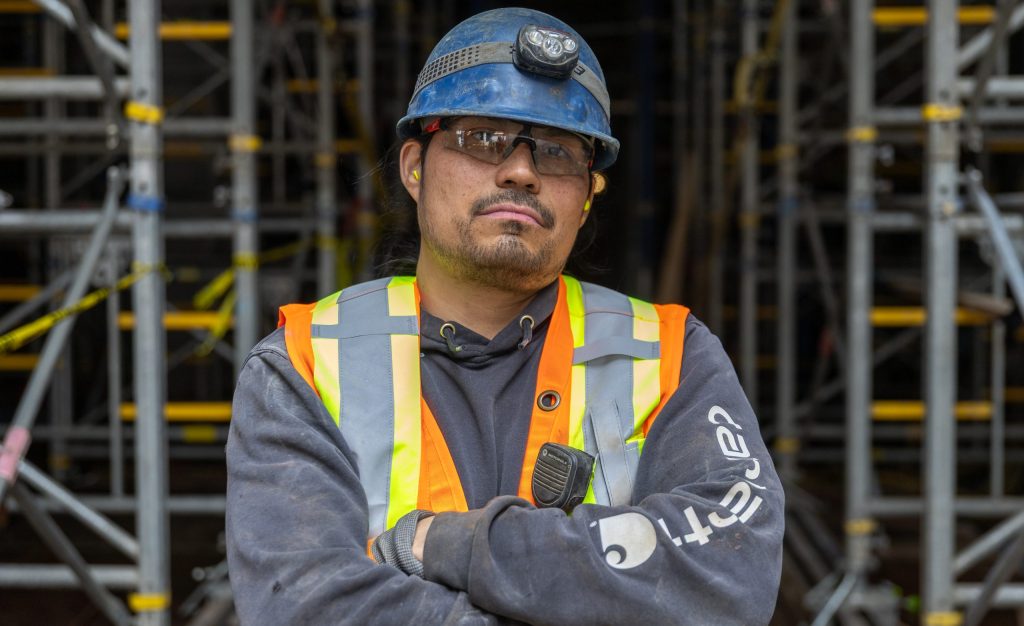 Picture off a construction worker looking at the camera, hands crossed, with scaffolding behind him.